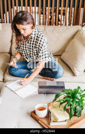 Woman with long brown hair sitting on a sofa with a laptop computer and notebook, working. Stock Photo