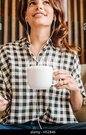 Woman with long brown hair, wearing headphones and chequered shirt, holding a mug. Stock Photo