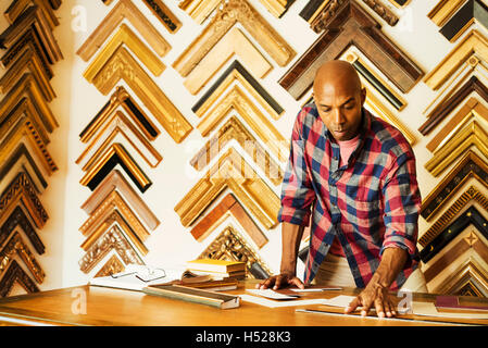 Man working at a picture framers, a large selection of frames on the walls. Stock Photo