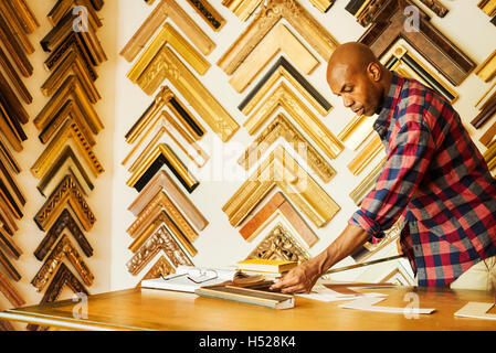 Man working at a picture framers, a large selection of frames on the walls. Stock Photo