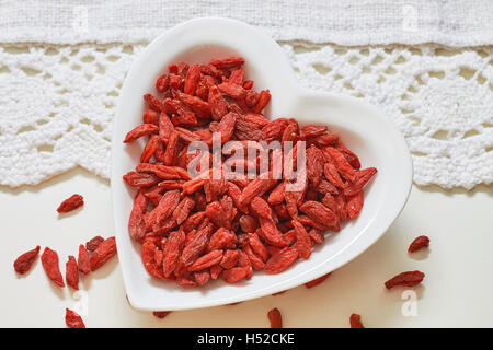 Dried goji berries in heart shaped ceramic bowl on white table. Top view Stock Photo