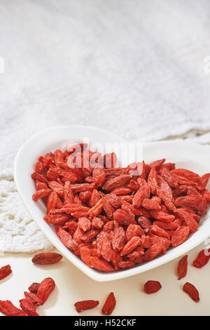 Dried goji berries in heart shaped ceramic bowl on white table. Copy space Stock Photo