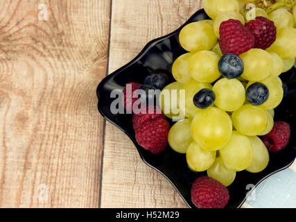 A photo close up grapes, raspberries and blueberries on a black saucer standing on a wooden table. Copy space on the left side.F Stock Photo