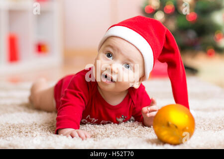 Funny baby wearing Santa hat and suit. Kid boy lying on tummy in front of Christmas tree at home Stock Photo