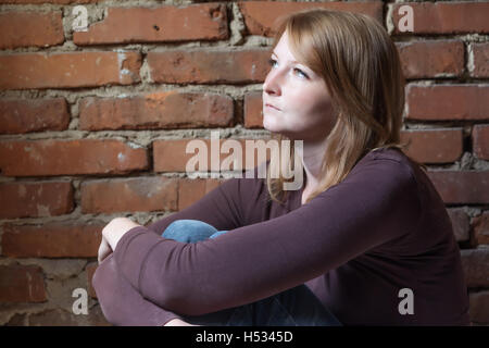 Side view of a young woman, who is sadly sitting in front of a brick wall and is looking to natural light. Stock Photo