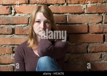 Portrait of pensive young woman sitting in front of brick wall in natural light. Woman is looking at the camera. Stock Photo