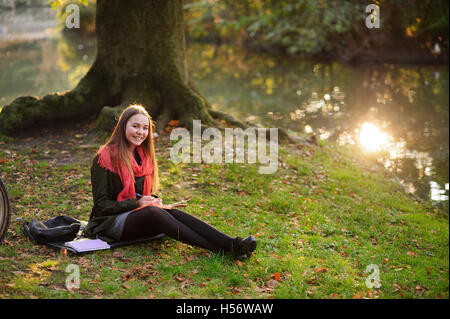 Picturesque autumn park. A girl in a bright red scarf sits on the ground, strewn with small autumn leaves. She writes something Stock Photo