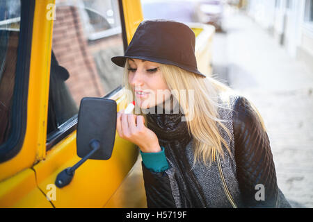 Sweet young woman applies red lipstick looking at the car mirror on a sunny autumn day Stock Photo