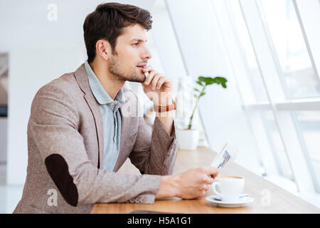 Thoughtful young businessman holding newspaper while having coffee break indoors Stock Photo