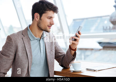 Young casual man in jacket texting message on smartphone while sitting indoors Stock Photo