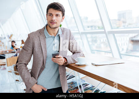 Handsome young man in jacket holding mobile phone while having lunch break Stock Photo