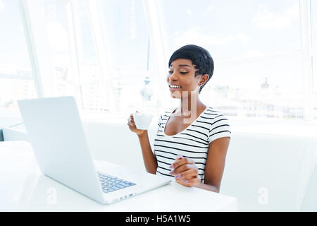 Smiling african american young businesswoman sitting at workplace and drinking coffee Stock Photo