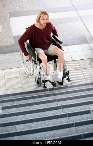 young man sitting in wheel chair in front of stairs and looking sad Stock Photo
