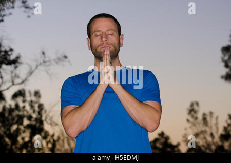 Man praying alone at dusk in an outdoor park wearing a blue t-shirt with shades of the sunset in the background. Stock Photo
