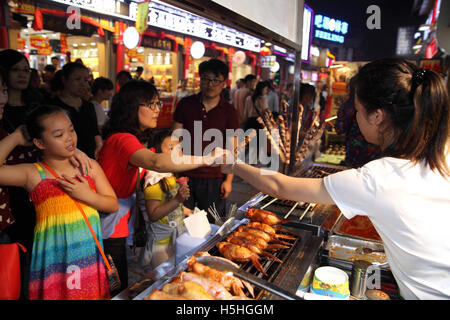 A Chinese mother buys chicken kebab on skewers for her daughters from a woman at a food stand.   The market street, Yangshuo Stock Photo