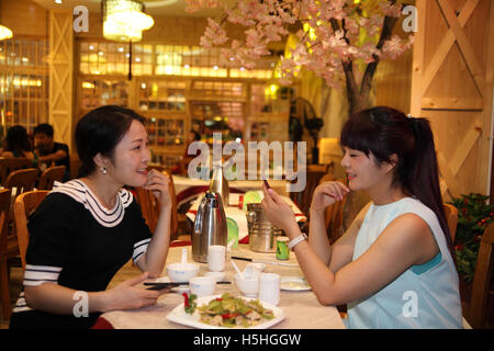 Relating in modern age, young Chinese women eat in a restaurant, talk and smile while one of them reads messages on her smartphone. Yangshuo, China. Stock Photo