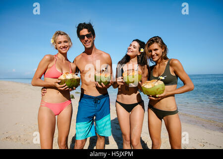 Portrait of young man and women in swimsuits holding fresh coconuts by the sea. Group of young friends on summer beach vacation. Stock Photo