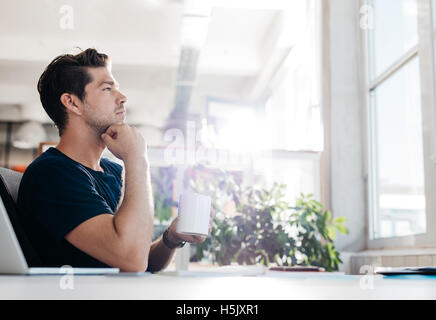 Side view shot of young businessman with coffee in hand sitting at his desk. Man sitting in office and thinking. Stock Photo