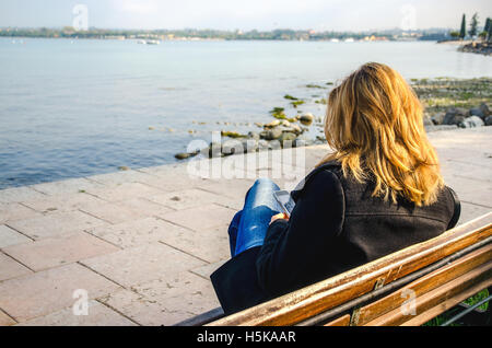blonde woman seen from behind using smartphone on the lake shore Stock Photo