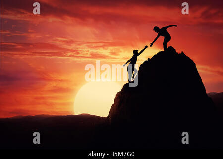 Hikers climbing on rock, mountain at sunset, one of them giving hand and helping to climb. Friendly assistance and support  in a Stock Photo