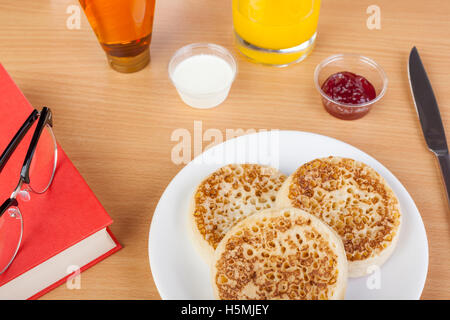 Fresh crumpets on a plate with a book and reading glasses aside strawberry jam and cream Stock Photo