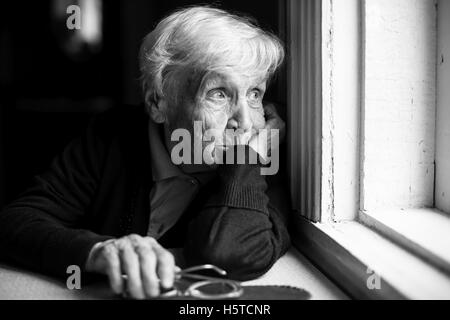 An elderly woman sadly looking out the window, a black-and-white photo. Stock Photo