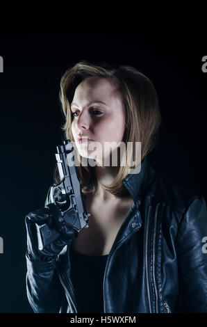 Blonde woman in black leather jacket holding a gun in the dark Stock Photo