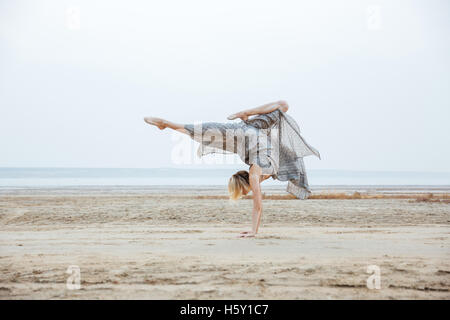 Beautiful young fitness woman in dress standing on arms and posing on the beach Stock Photo