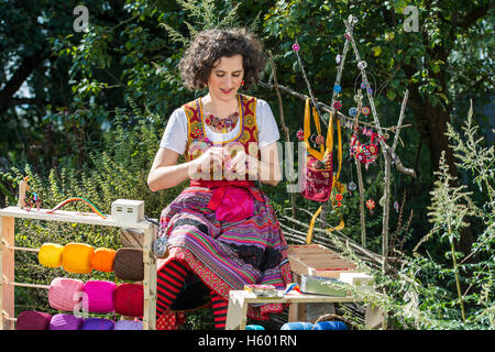 Button maker, young woman in colorful dirndl with Posamentenknopf Collier siting in garden and working on Posamentenknopf Stock Photo