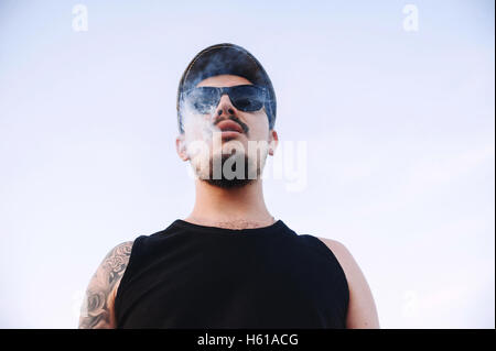 Close-up, portrait of young guy wearing a cap sunglasses with beard and mustache in tattoos, exhaling smoke from cigarette Stock Photo