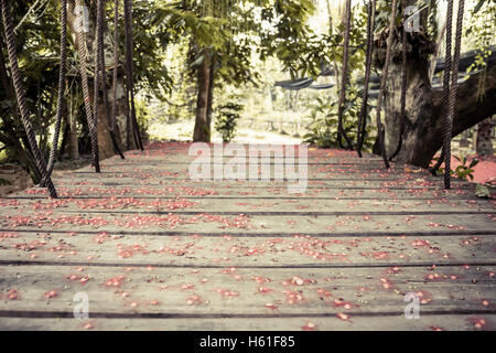 Old wooden suspension bridge with ropes in tropical forest covered with red flowers with selective focus on wooden planks Stock Photo