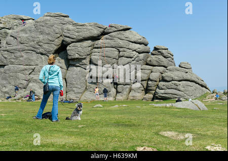 Woman walker with dog on lead stops to watch rock climbers attempt to climb Haytor Tor on Dartmoor in Devon UK Stock Photo