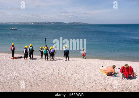 Young people learn to canoe on a beach in Brixham, Devon with Torquay in the background whilst a relaxing couple look on. Stock Photo