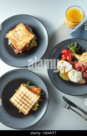 Breakfast with sandwiches on plates, benedict eggs with tomatoes and ham with cutlery and fresh orange juice Stock Photo