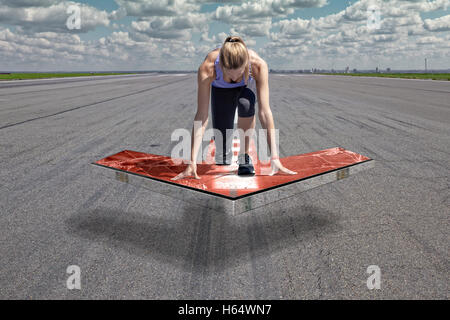 Female runner kneels in start position on a red floating arrow platform, which is placed above an airport runway surface. Stock Photo