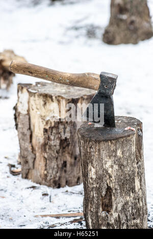 Ax stuck in log of wood on the background of the winter forest and huts. Stock Photo