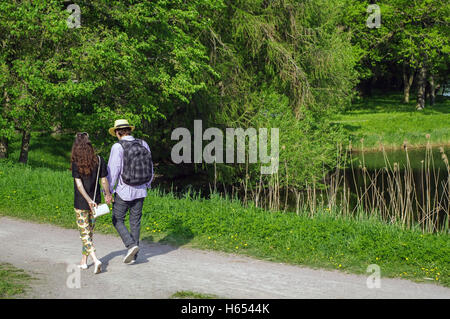 a happy young couple walking in summer park holding hands with a pond on side Stock Photo