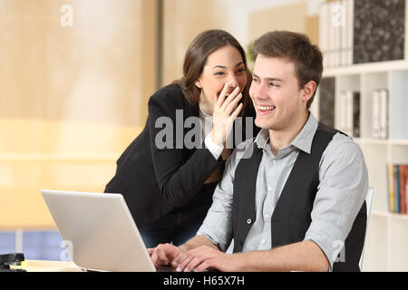 Gossip businesswoman telling secrets to the ear of a businessman sitting on a desktop at office Stock Photo