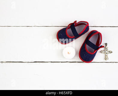 Pair of navy and red baby booties, candle and cross on white wood table Stock Photo