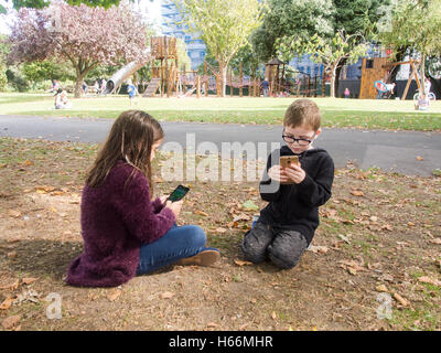 two children play games on mobile phones in a public park with a play area behind them. Stock Photo