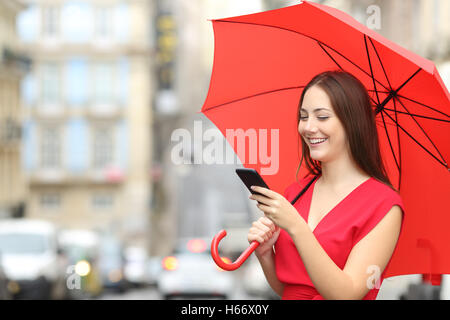 Portrait of a happy woman wearing red blouse texting on a smart phone under an umbrella in a rainy day Stock Photo