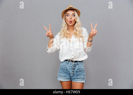 Funny excited young woman showing peace sign over gray background Stock Photo