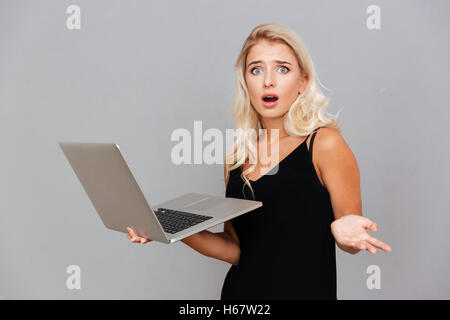 Amazed frustrated woman in black dress holding laptop over gray background Stock Photo