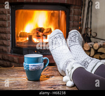Warming and relaxing near fireplace. Woman feet near the cup of hot drink in front of fire. Stock Photo