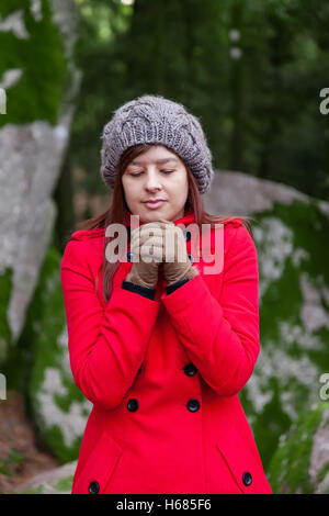 Young woman shivering with cold on a forest wearing a red overcoat, a beanie and gloves during winter Stock Photo