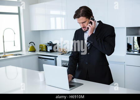 Businessman using laptop while talking on mobile phone in kitchen Stock Photo