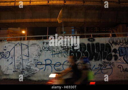 A couple ride on a scooter at night in front of a wall covered with a graffiti reading 'Free Oxygen' and '2014 Bangkok' in Bangkok, Thailand. Stock Photo