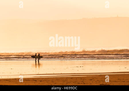 Early morning sunlight silhouetting two surfers standing on Gwithian Towans beach in Cornwall. Stock Photo