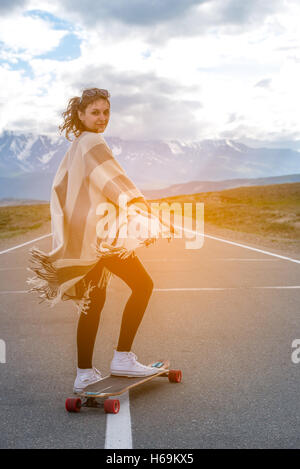Young cute girl rides skateboard on road in the mountains Stock Photo