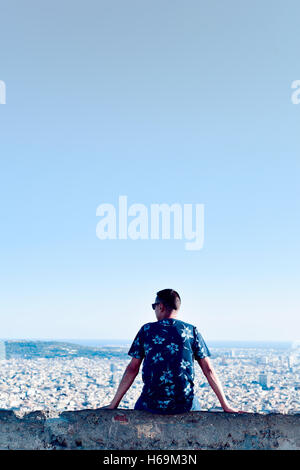 a young caucasian man, seen from behind, sitting at the top of a hill observing the city below him Stock Photo